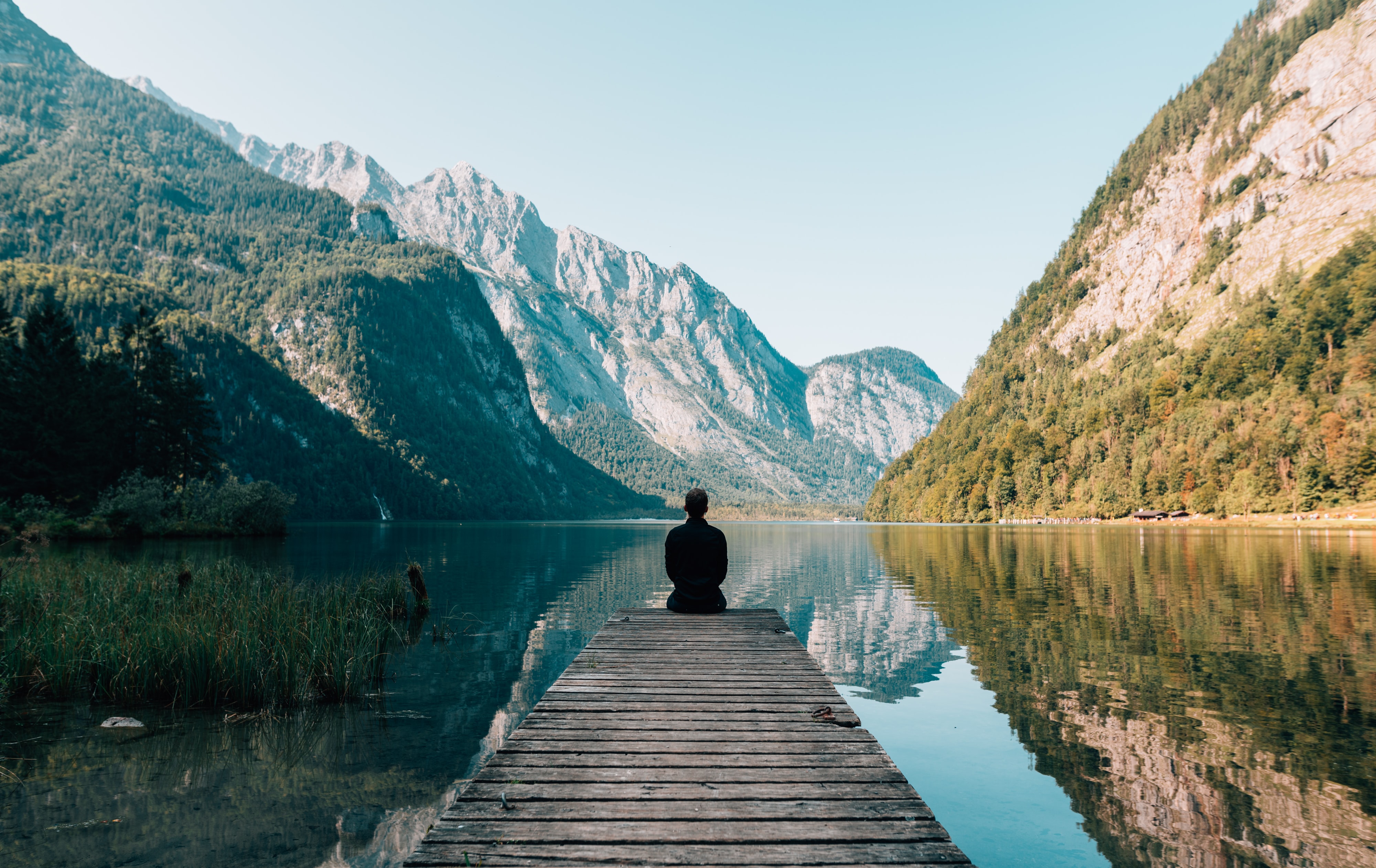 person sitting at the end of a pier that leads out to a clear lake with back to the camera. Mountains with some growth in the background.